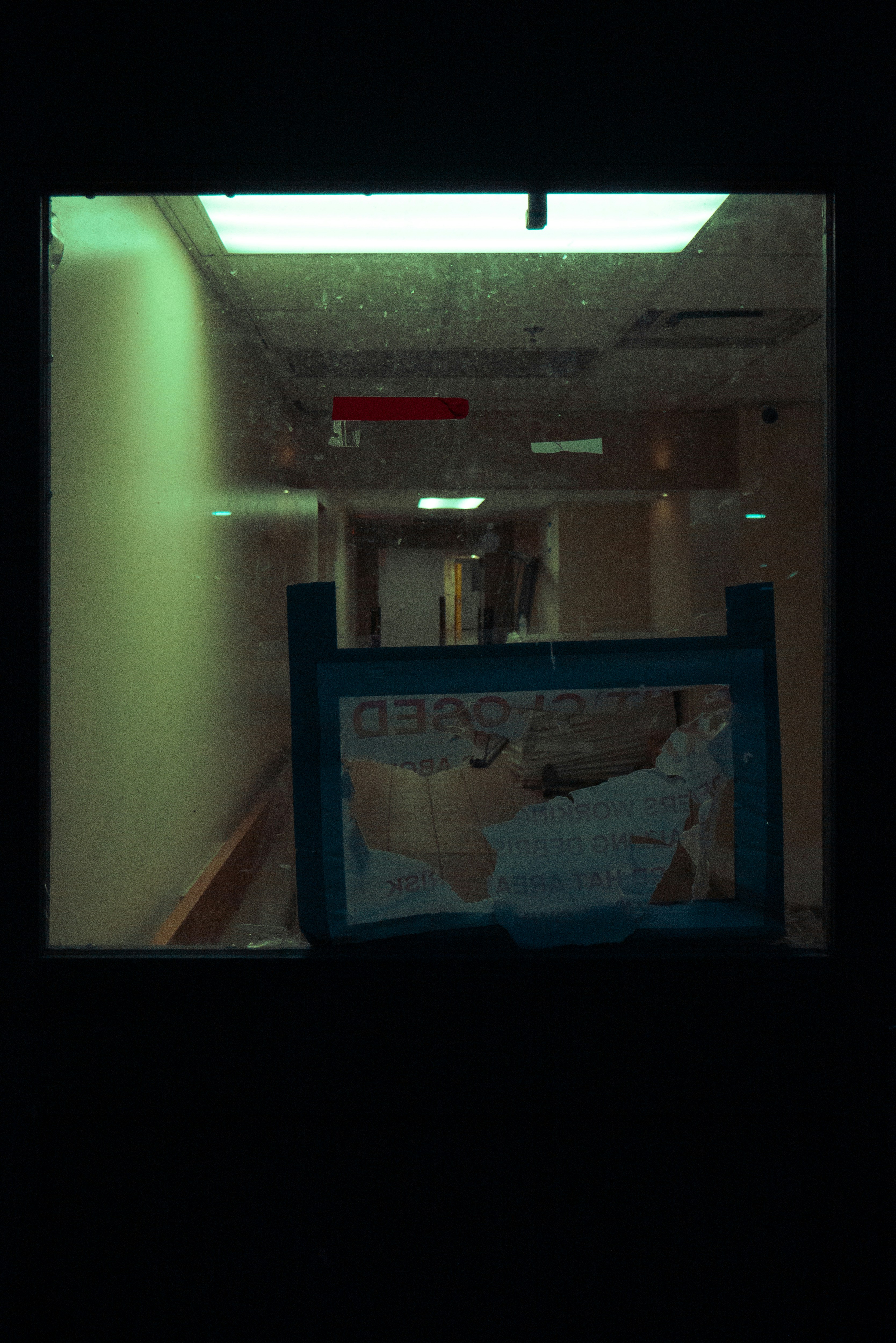a dark and somewhat blurry photo, taken through a window, of a closed business. the word "closed" is visible on a sign in the window, but the rest of the sign is torn and illegible. the hallway beyond the window is dimly lit with fluorescent lighting.  the overall mood is one of emptiness and abandonment.