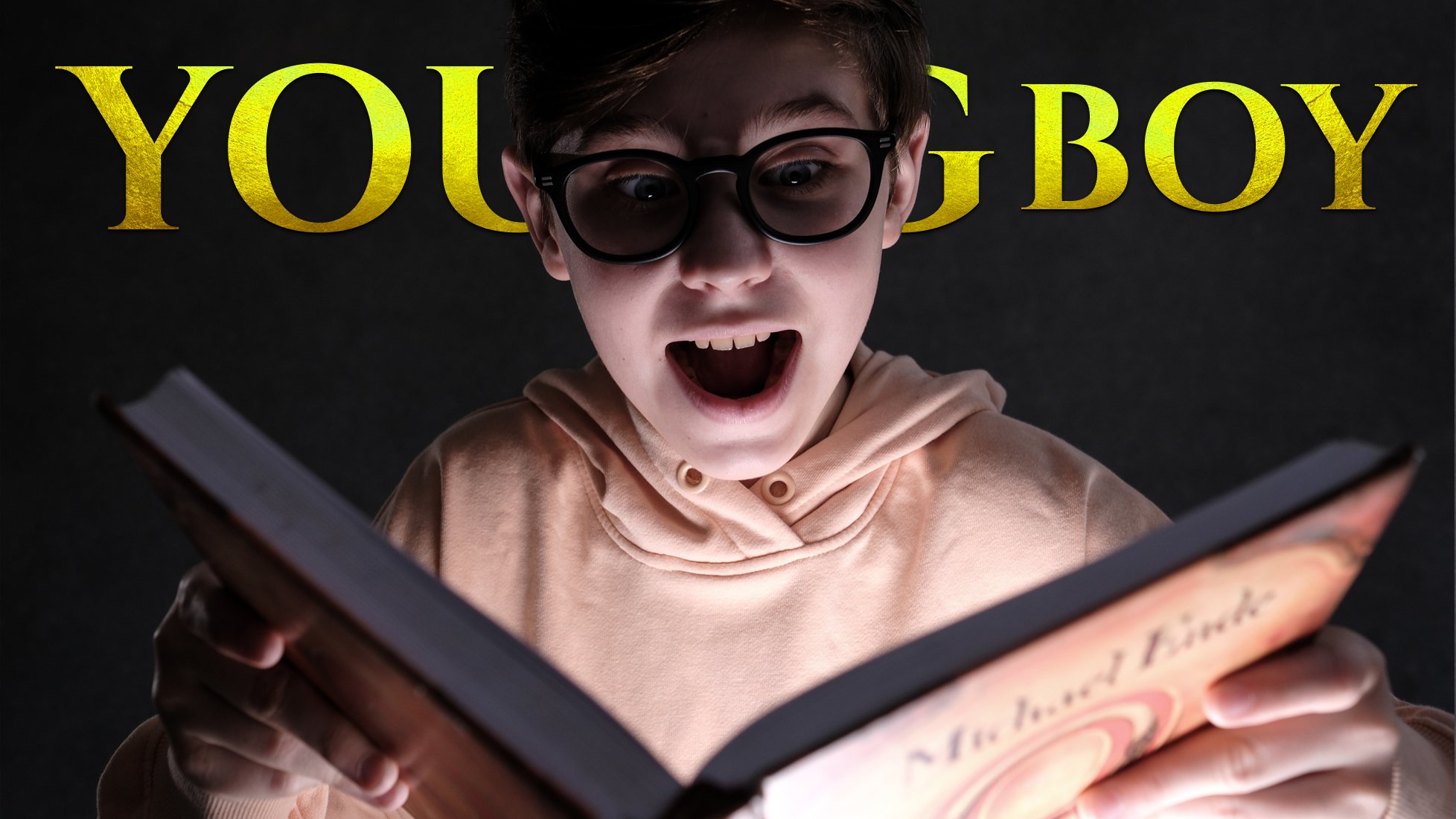 a closeup shot of a young boy with glasses, wearing a peachcolored hoodie, looking surprised and openmouthed as he reads a book. the book is illuminated, casting a glow on his face. the background is dark, and the words "young boy" are written in gold lettering above him. the overall mood is one of excitement or surprise.