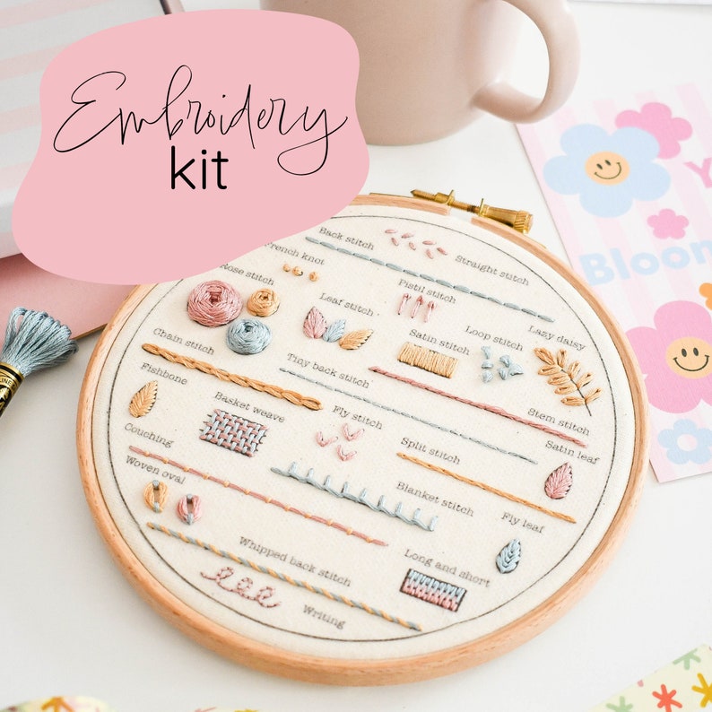 a flat lay photograph of a beginner embroidery kit. the kit features an embroidery hoop with various stitches sewn onto it as a sampler guide. the stitches are labeled with their names and worked in pastelcolored embroidery floss.  the hoop rests on a white surface, and a cup and a sheet of paper with a floral design peek in from the top right and bottom left corners respectively, creating a bright and inviting crafting scene.