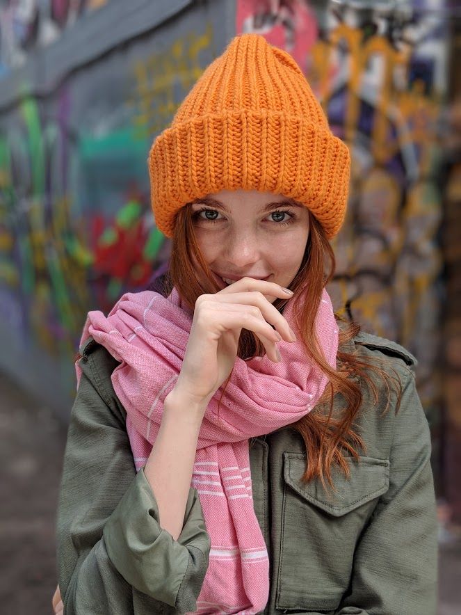 a woman with red hair wearing an orange beanie and a pink scarf smiles at the camera. she is standing in front of a graffiti wall.