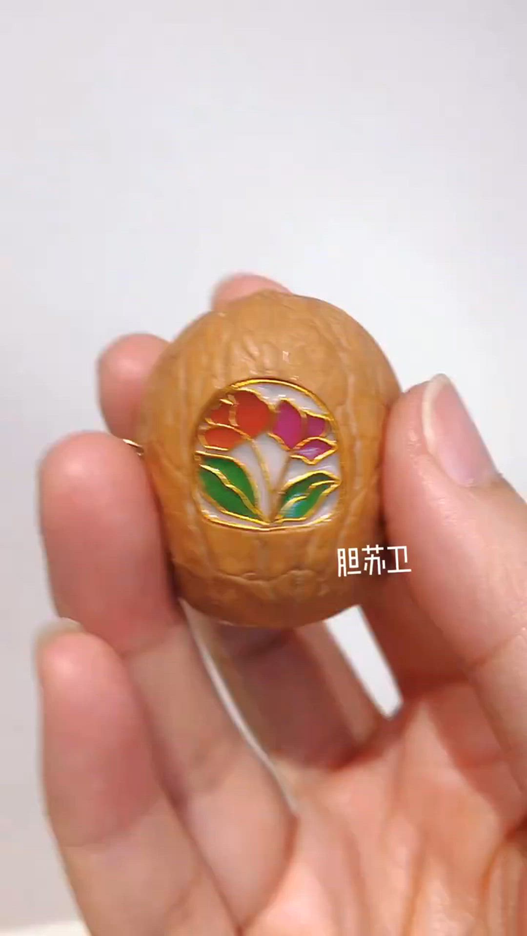 a hand is holding a walnut shell with a detailed flower ornament on the shell.  the flower has red and gold details and is against a white background.