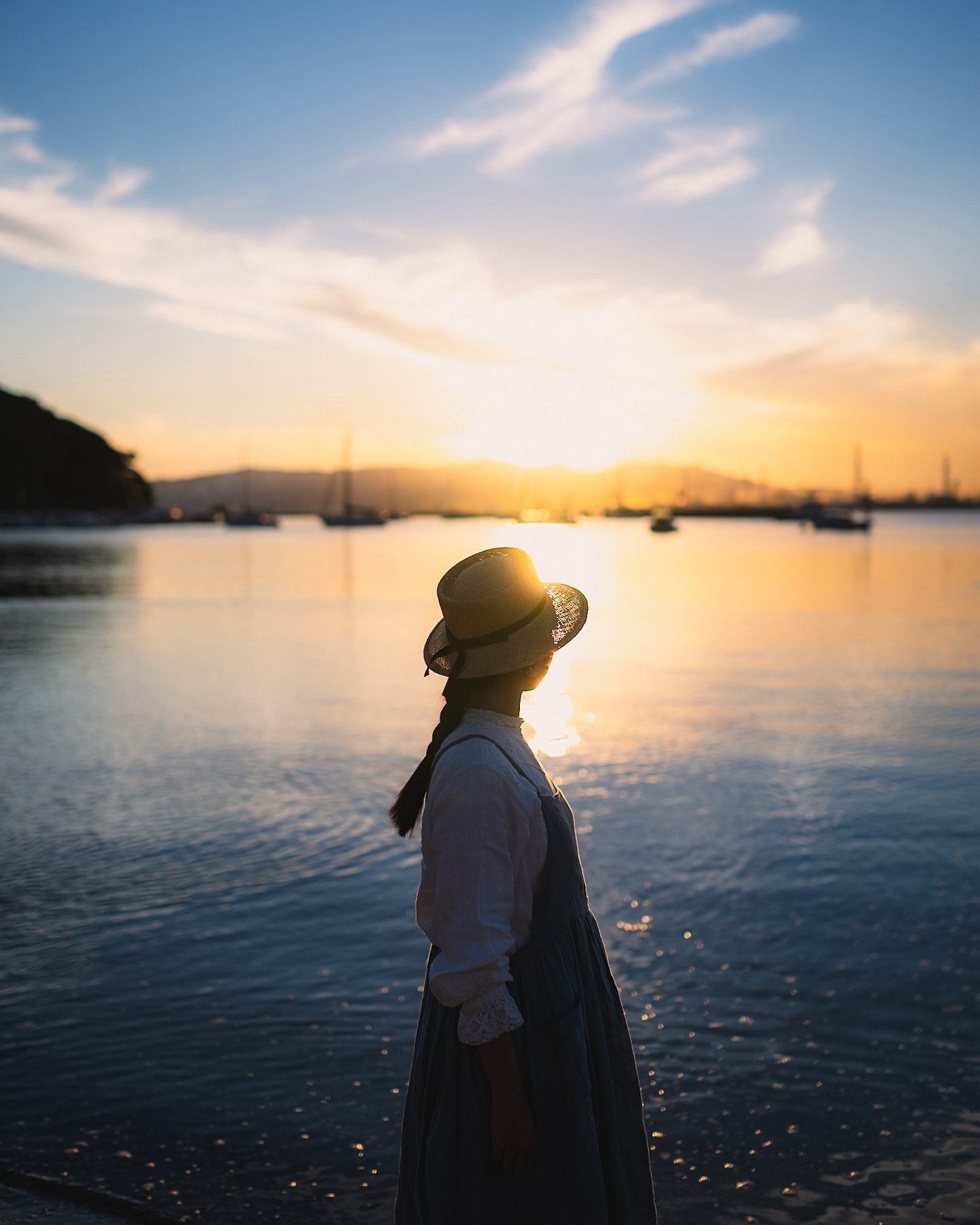 a silhouette of a woman in a dress and sun hat stands on the shore of a calm ocean at sunset. the sun is setting behind her, creating a warm golden light. the water is calm, and there are boats visible in the distance. the sky is a mix of blue and orange hues. the overall mood is peaceful and serene.