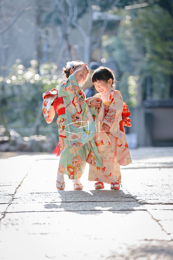 two young girls, dressed in traditional japanese kimonos, are walking along a stone path at a temple. the girls are smiling and appear to be enjoying themselves. the scene is bathed in warm sunlight, creating a cheerful and festive atmosphere.