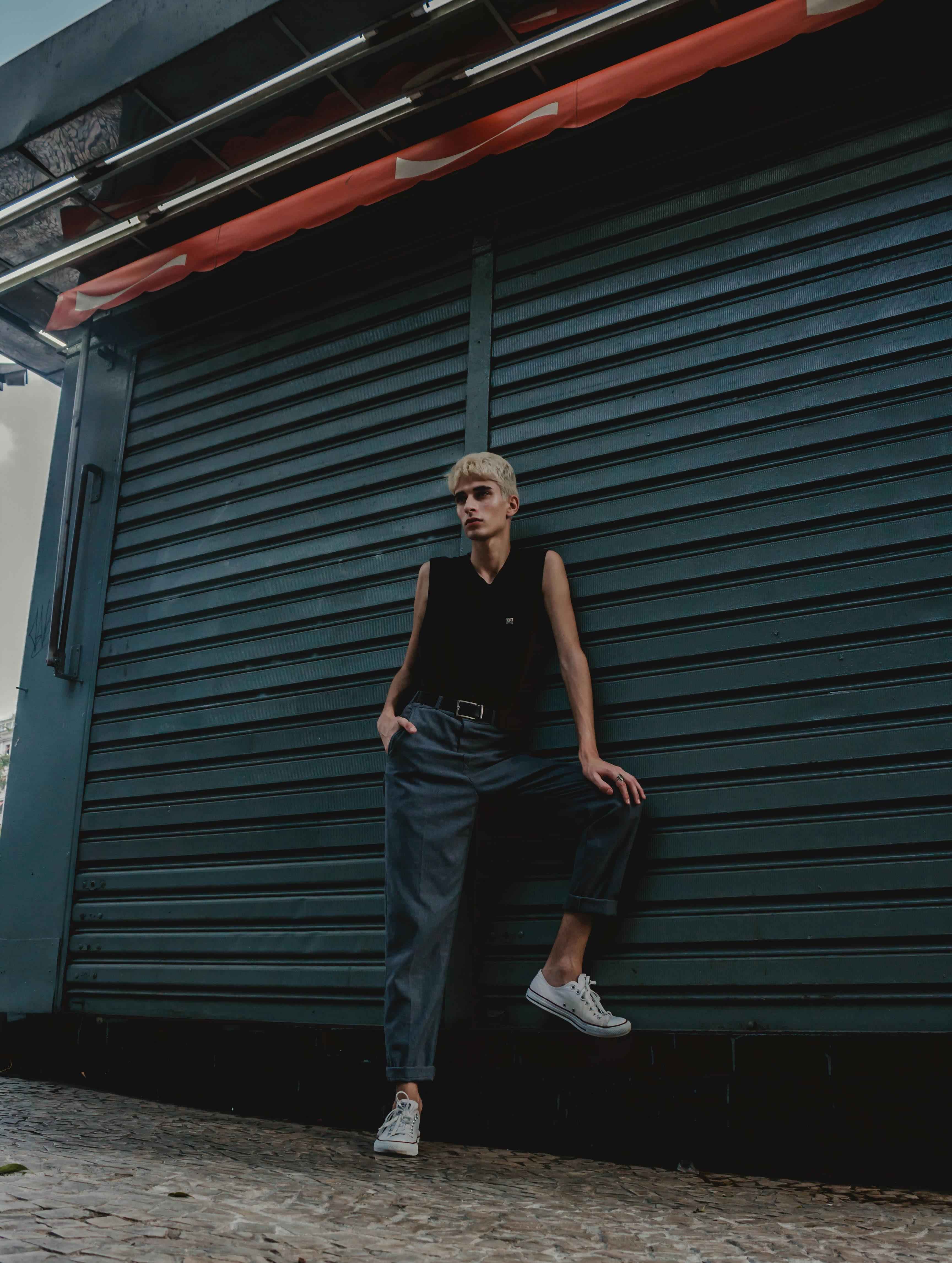a man with short blonde hair is standing in front of a closed dark teal metal shutter. he is wearing a black sleeveless top, dark blue trousers, a black belt, and white sneakers. he has one hand in his pocket and is leaning slightly against the shutter. the scene appears to be in an urban environment.