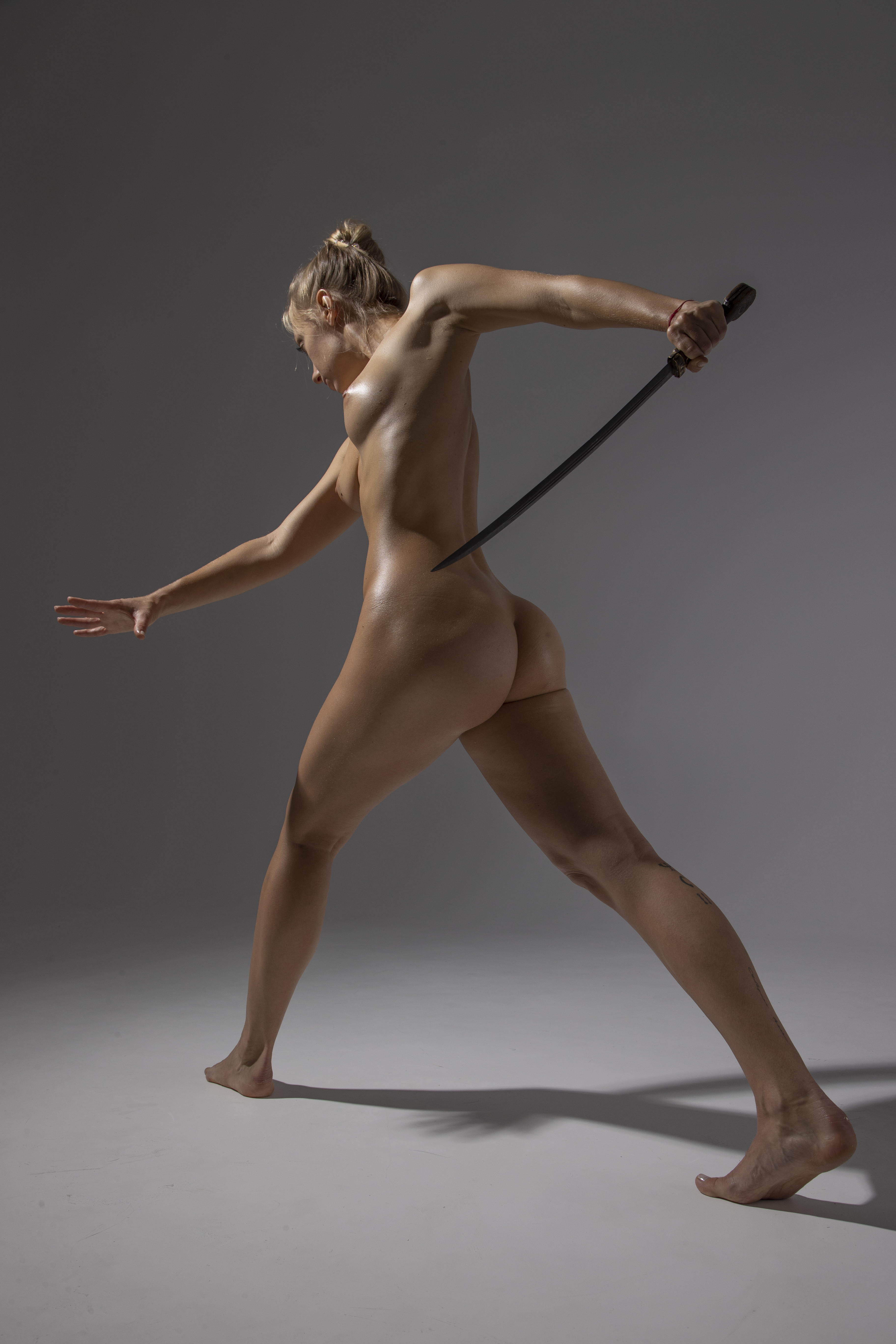 a nude woman stands in a dynamic pose with a sword in her hand. her body is athletic and toned, showcasing her muscles. she is positioned against a gray background in a studio shot. the full shot captures her strength and warriorlike stance.