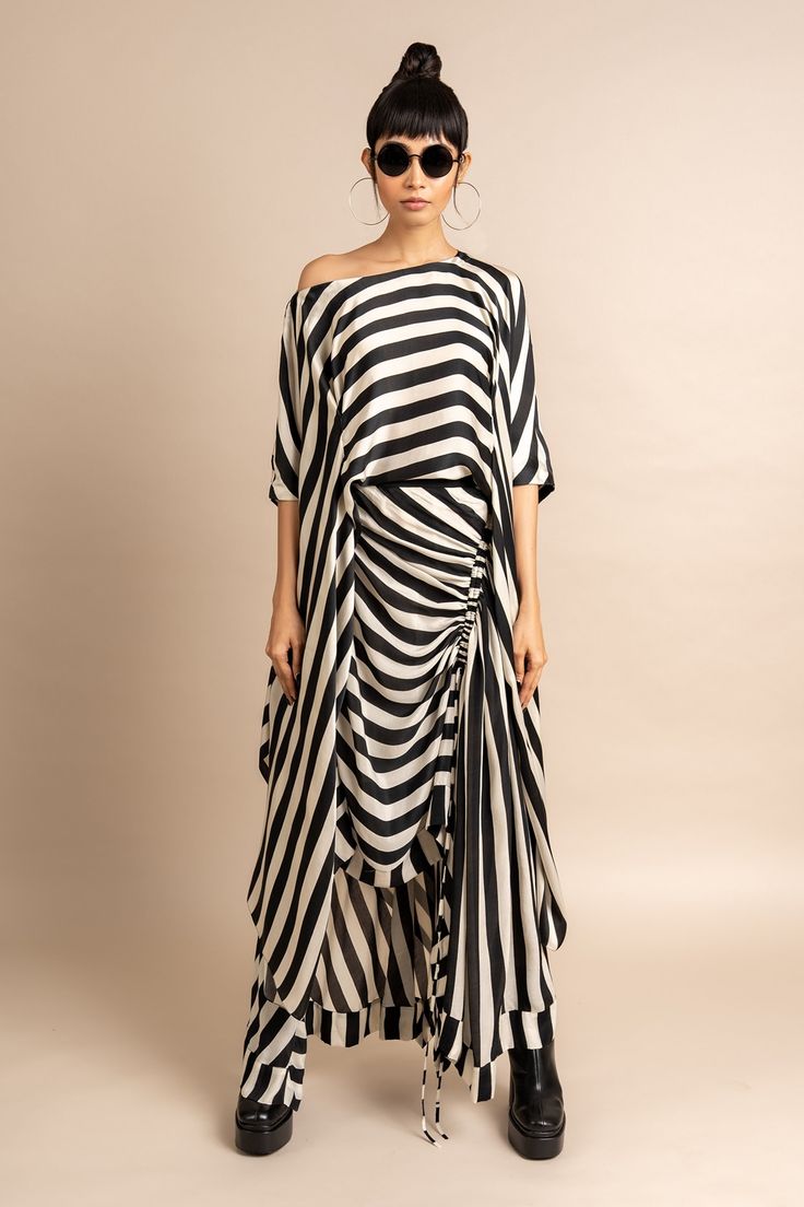a woman is standing in front of a beige background, she is wearing a long black and white striped dress and black boots. the dress is cinched in at the waist and has a gathered skirt. she is also wearing sunglasses and has her hair pulled back in a ponytail. the woman is looking at the camera with a serious expression.