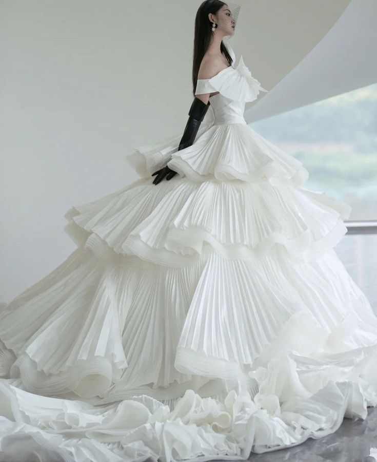 a model wearing a white dress with multiple layers of ruffles and pleats. the dress is strapless and has a fitted bodice. the model is also wearing black gloves and has her hair pulled back. the image is taken in a studio setting. the background is white and blurred, which helps to emphasize the dress.  the model looks elegant and sophisticated in the dress, which is perfect for a formal event.