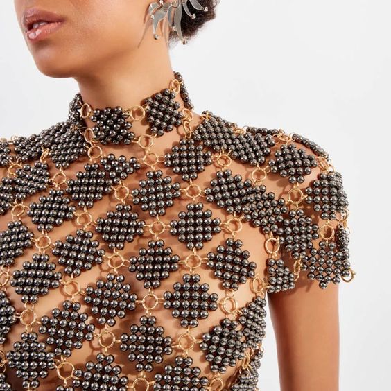 a closeup shot shows a woman wearing a unique piece of jewelry, which resembles a shirt or top made from black beads arranged in diamond patterns, connected by gold rings. the beaded pattern extends to a high choker around her neck. she is also wearing dangling silver earrings. the jewelry is designed to reveal sections of bare skin underneath.