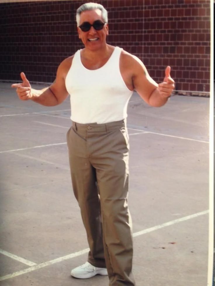 a middleaged man wearing sunglasses, a white tank top, and khaki pants stands on a concrete surface in front of a brick wall, giving a thumbs up with both hands and smiling. he is also wearing white sneakers.
