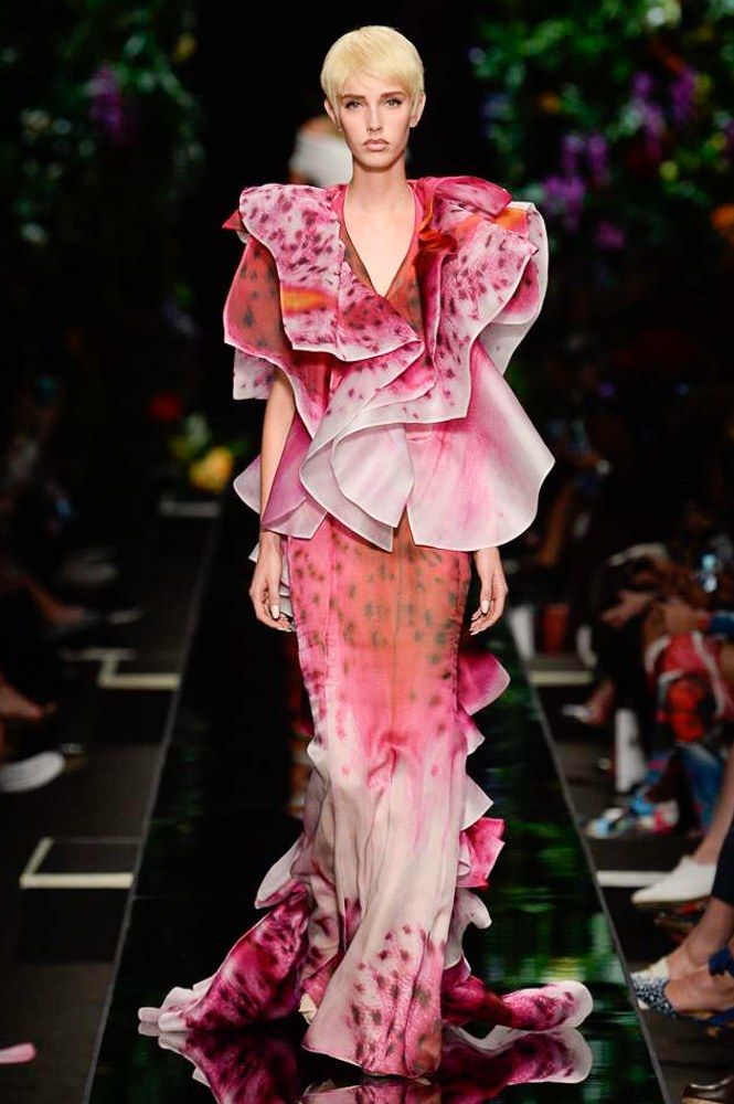 a fashion model walks the runway in a pink and floral gown. the dress is a stunning work of art with delicate layers of fabric and a beautiful floral print. the model's hair is styled in a sleek bob and her makeup is natural and glowy. she is wearing a pair of strappy heels that compliment the dress perfectly.