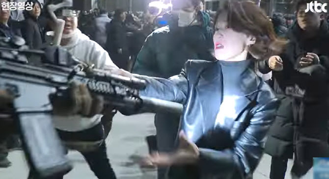 a woman in a leather jacket appears to be in a crowd, with at least one person holding a gun in the foreground. the scene seems intense, with multiple individuals present in what looks like an outdoor setting. there is a firearm pointed towards the camera.
