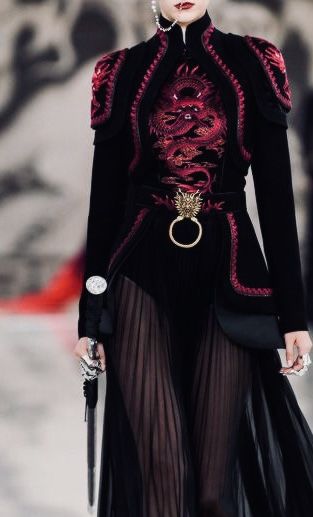 a woman is wearing an elaborate black dress with red dragon embroidery. the dress has a high collar, shoulder pads, and a belt with a large ring. the bottom of the dress is made of sheer pleated fabric. the woman is holding a sword and is wearing a ring. she appears to be walking on a runway.