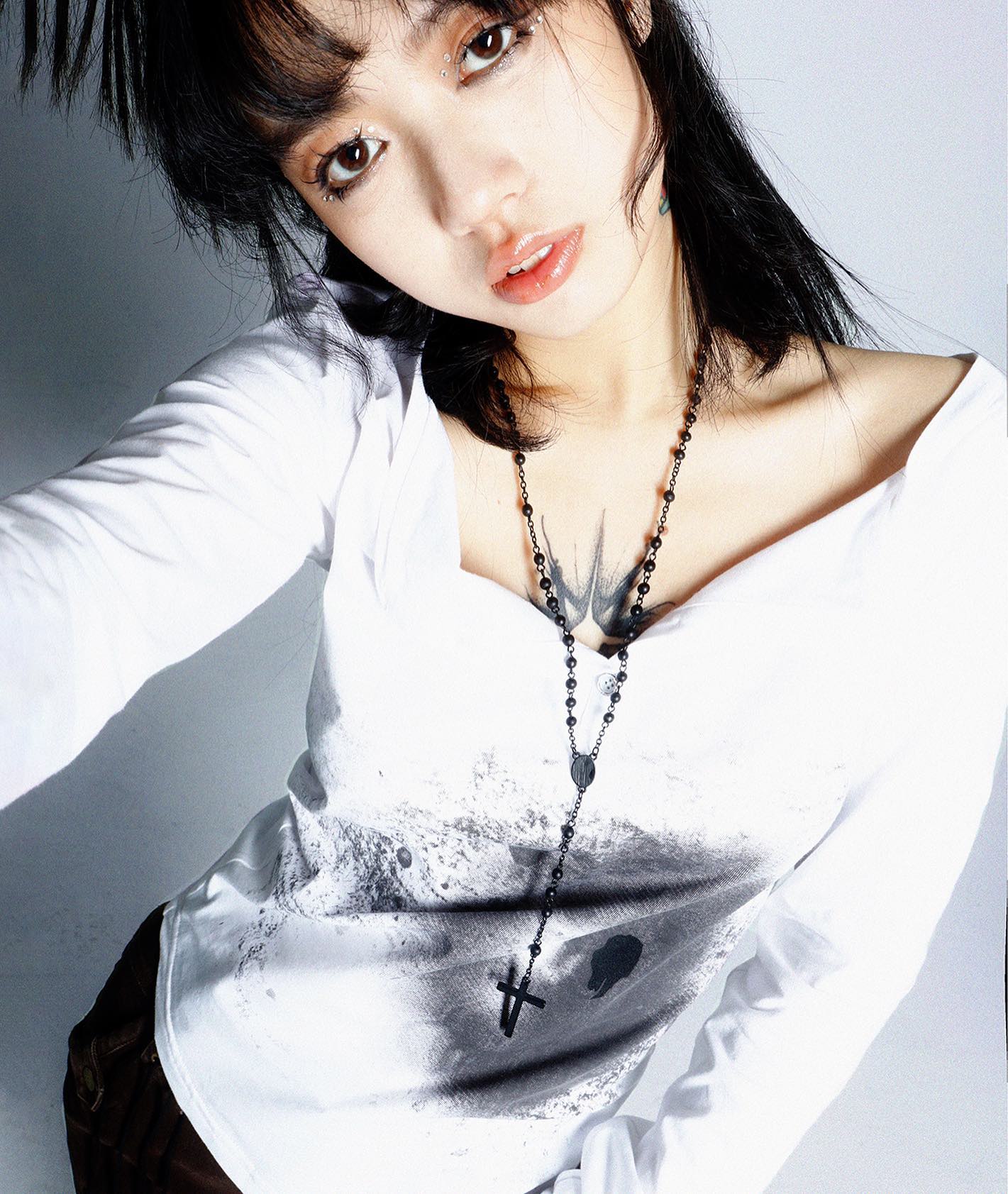 closeup shot of a young asian woman with dark hair and bangs, wearing a white longsleeved shirt with a vneck and a black rosary necklace. she has a tattoo on her chest and is looking at the camera. the image is styled in a fashionforward manner with an edgy and cool aesthetic. the background is white.