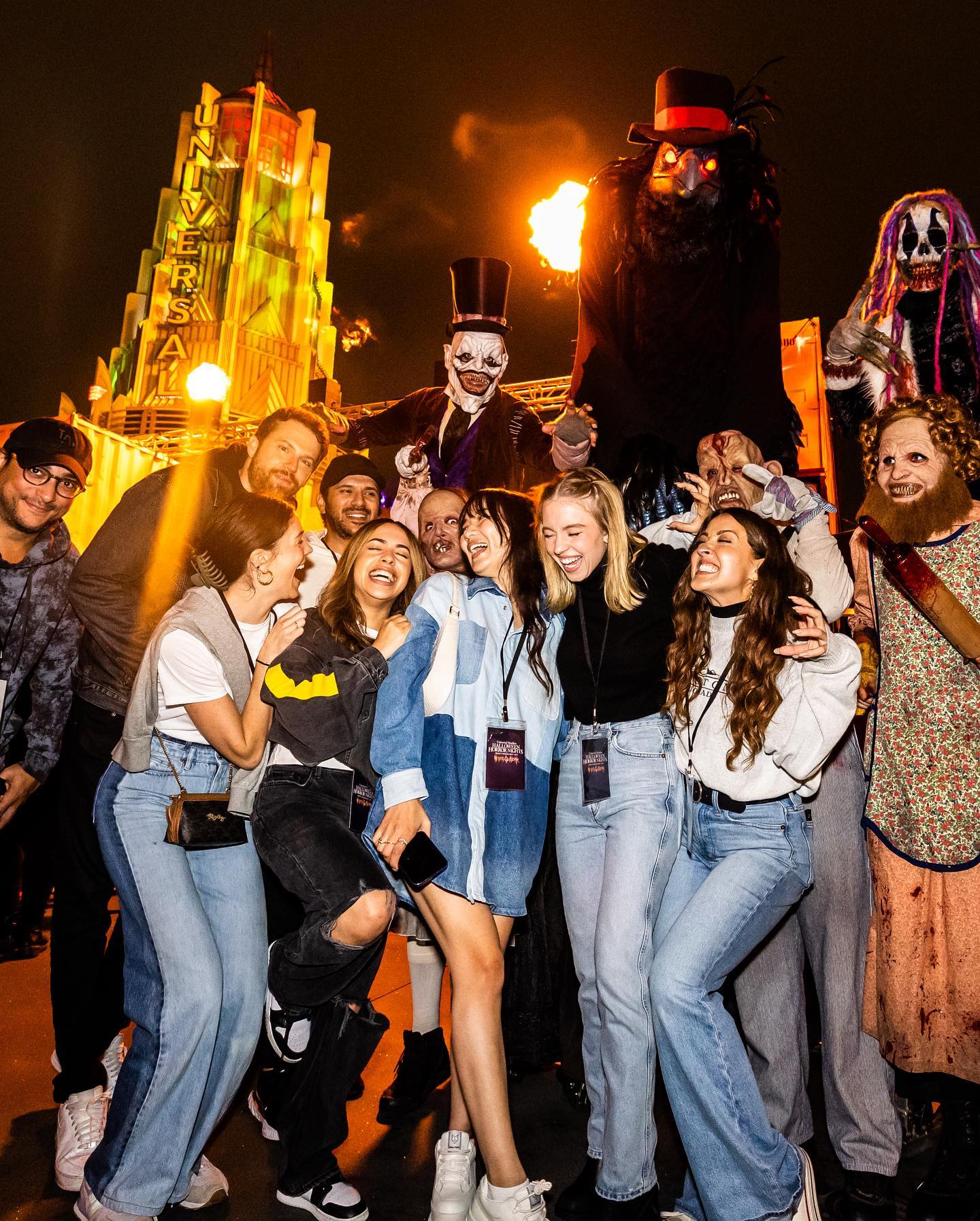 the image captures a large group of smiling people posing for a photograph at universal studios hollywood's halloween horror nights.  they're clustered around several scare actors in elaborate costumes; one is a tall, imposing figure resembling a raven or crow, another is a tophatted jesterlike character, and a third is a terrifying figure with long, dreadlocked hair.  the background features the iconic universal studios hollywood building, illuminated in colorful lights.  flames and other special effects are visible, adding to the atmosphere.  the overall scene is festive and energetic, highlighting the exciting and fun aspects of the halloween event.  the group is primarily comprised of young adults, many wearing casual jeans and jackets. the photo seems to have been taken by a professional photographer given the lighting and composition.