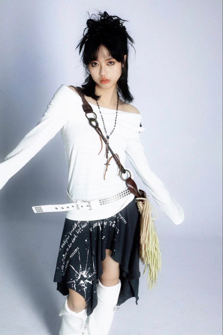 a young woman with dark, choppy hair is posing in a studio. she's wearing a white offtheshoulder longsleeved top, a black asymmetrical mini skirt with white text and graphic detailing, a white studded belt, and white thighhigh boots. she carries a brown leather crossbody bag with a fringe detail.  the overall style is grunge or punkinspired, with a mix of textures and edgy accessories.  the background is plain white, focusing attention on the model and her outfit.