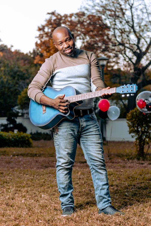a smiling black man with a bald head is standing outdoors in a park, playing a blue acoustic guitar. he's wearing a beige and grey sweater and light blue jeans. the background is slightly blurry, showing autumnal trees with brown and orange leaves. the grass is dry and brown. the man appears relaxed and happy.