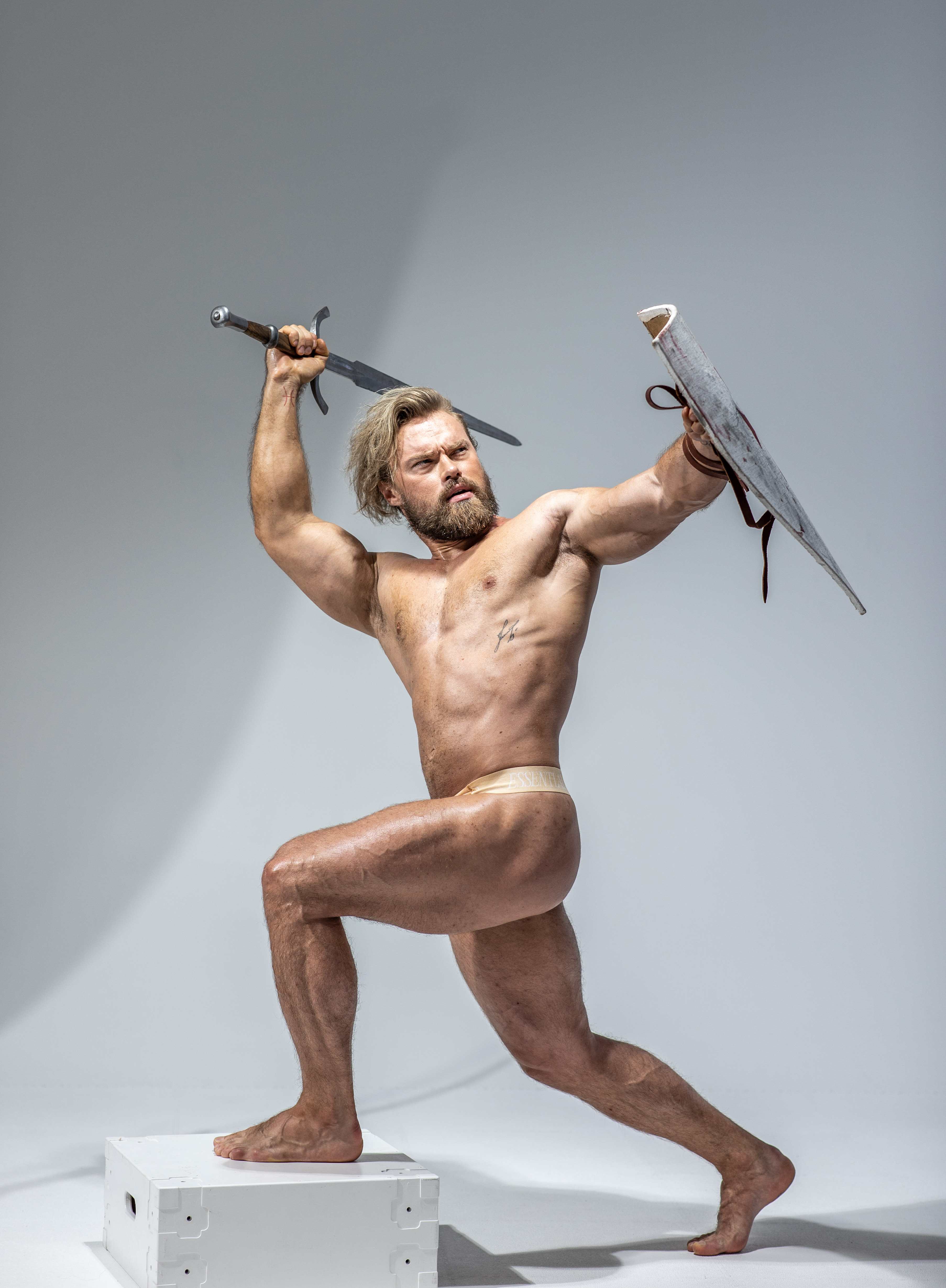 a muscular man with long blonde hair and beard is posed dynamically against a light grey background. he's holding a sword in his right hand and a shield in his left, as if in the midst of a battle. his attire is minimal, wearing only a loincloth. the lighting is dramatic, highlighting his physique.