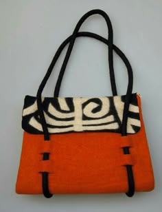 a close up image of a small orange handbag with black handles. the bag is made of felt and has a white and black design on the front. the bag is on a white surface.