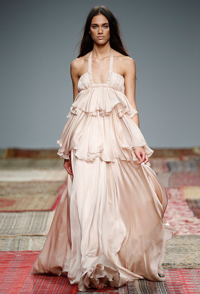 a young woman is on the runway, walking towards the camera. she is wearing a long, tiered, pale pink gown with ruffled detailing. her hair is long and dark, and she is looking forward with a serene expression.
