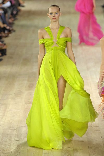 a model walks down a runway during a fashion show. she is wearing a lime green chiffon gown with a high slit. another model is partially visible wearing a hot pink gown. a group of people are seated in the background.