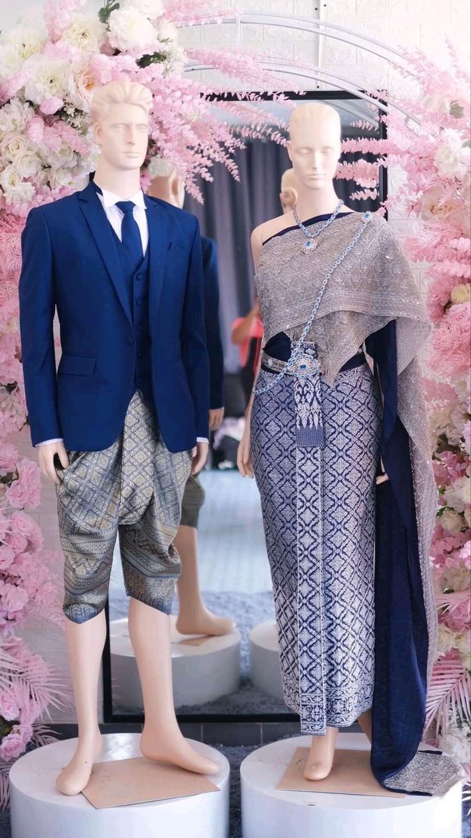 a bride and groom mannequin dressed in traditional thai wedding attire stand together in a floral setting. the bride is wearing a silver and blue dress with a white shawl. the groom is wearing a blue suit with a white shirt and tie. the mannequins are standing on white pedestals with flowers surrounding them. the flowers are pink and white. the bride and groom are looking at each other. the image is likely taken in a store or at a wedding venue.