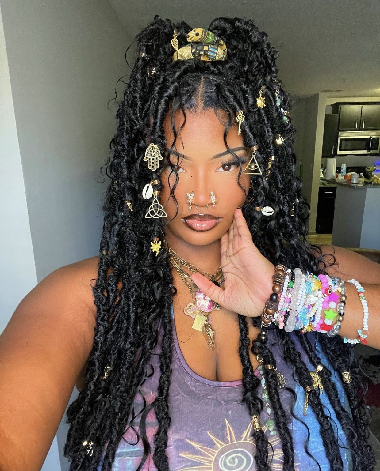 a young, confident, and beautiful black woman poses for a selfie.  she is wearing  long, black, curly braids that are accessorized with gold jewelry. she is also wearing several gold necklaces and a stack of bracelets on one wrist.  she is smiling slightly and her eyes are bright.