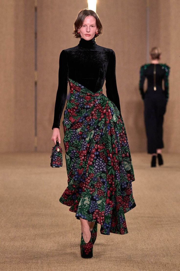 a runway model walks down the catwalk wearing a unique outfit. the outfit consists of a black velvet turtleneck top and a highlow skirt adorned with a vibrant fruit and berry print.  the skirt is draped and gathered, creating a layered and textural effect. the model carries a small, darkcolored handbag and is wearing darkcolored shoes with a pointed toe. the background is a neutral beige tone, drawing focus to the model’s outfit.  another model is visible in the background, out of focus. the overall impression is one of high fashion and elegant autumn/winter style.