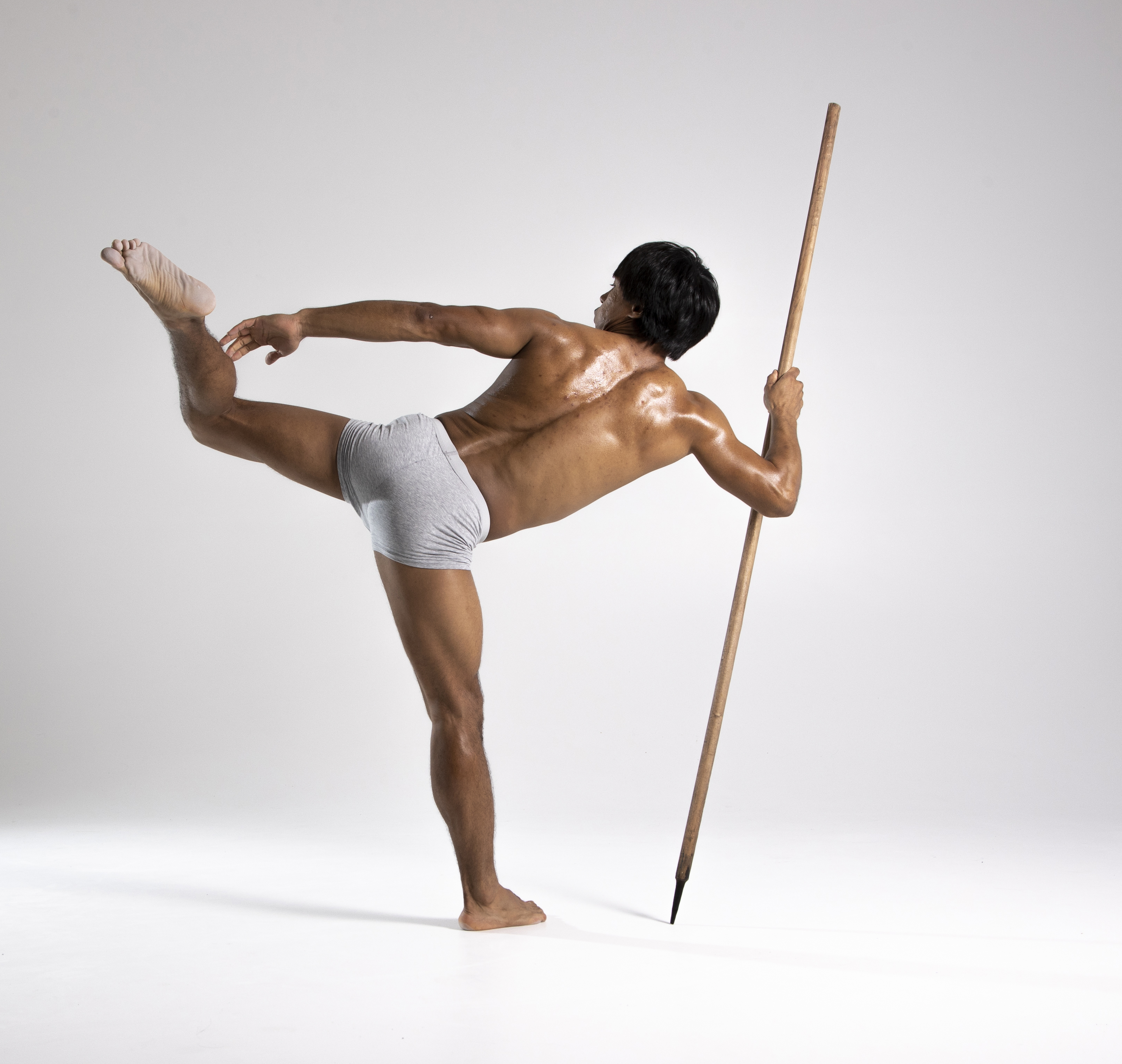 a muscular man is posing in a studio, balancing on one leg while holding a spear. he is wearing gray briefs and is barefoot. the background is a plain white. the man is displaying an athletic pose that combines yoga and contortion elements.