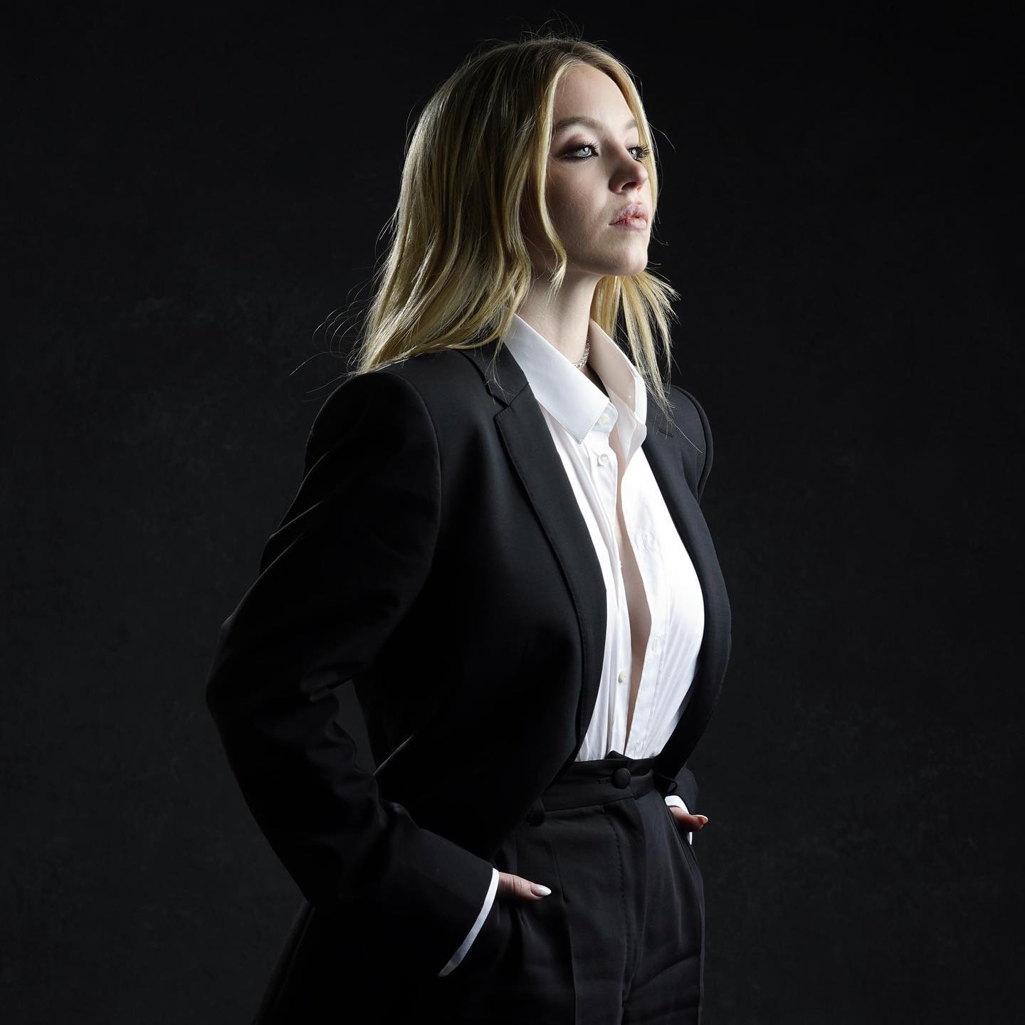 the image is a professional studio portrait of a blonde woman. she is wearing a black blazer over a crisp white shirt and black highwaisted trousers, creating a chic pantsuit look.  her hair is long and falls over her shoulders. she has a serious, perhaps pensive expression on her face and is looking off to the side, away from the camera. the background is dark and unblemished, drawing all focus to the woman.  the lighting highlights her features and the texture of her clothing. the overall style is sophisticated and suggests a confident, professional image, possibly for a promotional photoshoot or headshots.