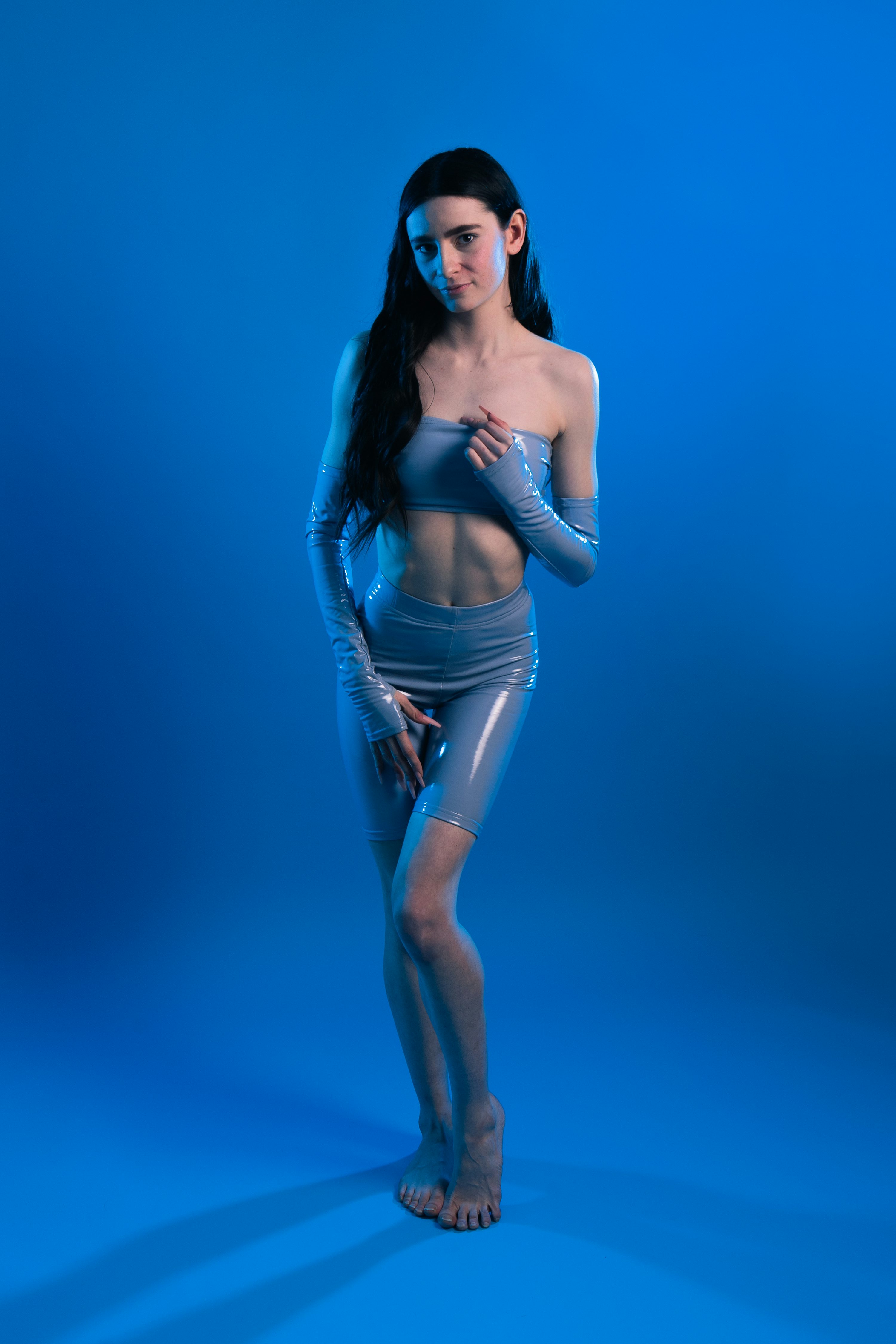 a young woman with long dark hair poses confidently in a sleek, light blue latex bodysuit and matching gloves against a vibrant blue background. the lighting creates a futuristic and stylish atmosphere. her pose is relaxed yet striking, showcasing the formfitting outfit and highlighting her physique. the overall aesthetic is minimalist and modern.