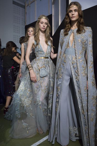 a captivating image showcasing three models gracefully adorned in exquisite, floorlength gowns. the dresses are predominantly blue with intricate silver embroidery, adding a touch of sparkle. each model exudes elegance, their attire enhanced by delicate jewelry including headbands. the central focus is on the two models in front, positioned on a runway. the overall setting implies a highfashion context, possibly a runway show or backstage area. the photograph expertly captures the models' poise and the meticulous detailing of their dresses, creating a scene that is both glamorous and fashionable.