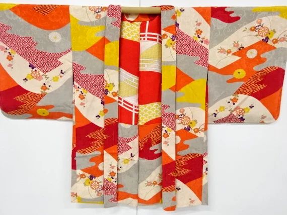 an image of a vibrant japanese kimono displayed on a hanger. the kimono features a colorful pattern with various geometric shapes and floral designs in shades of red, orange, yellow, gray, and white.