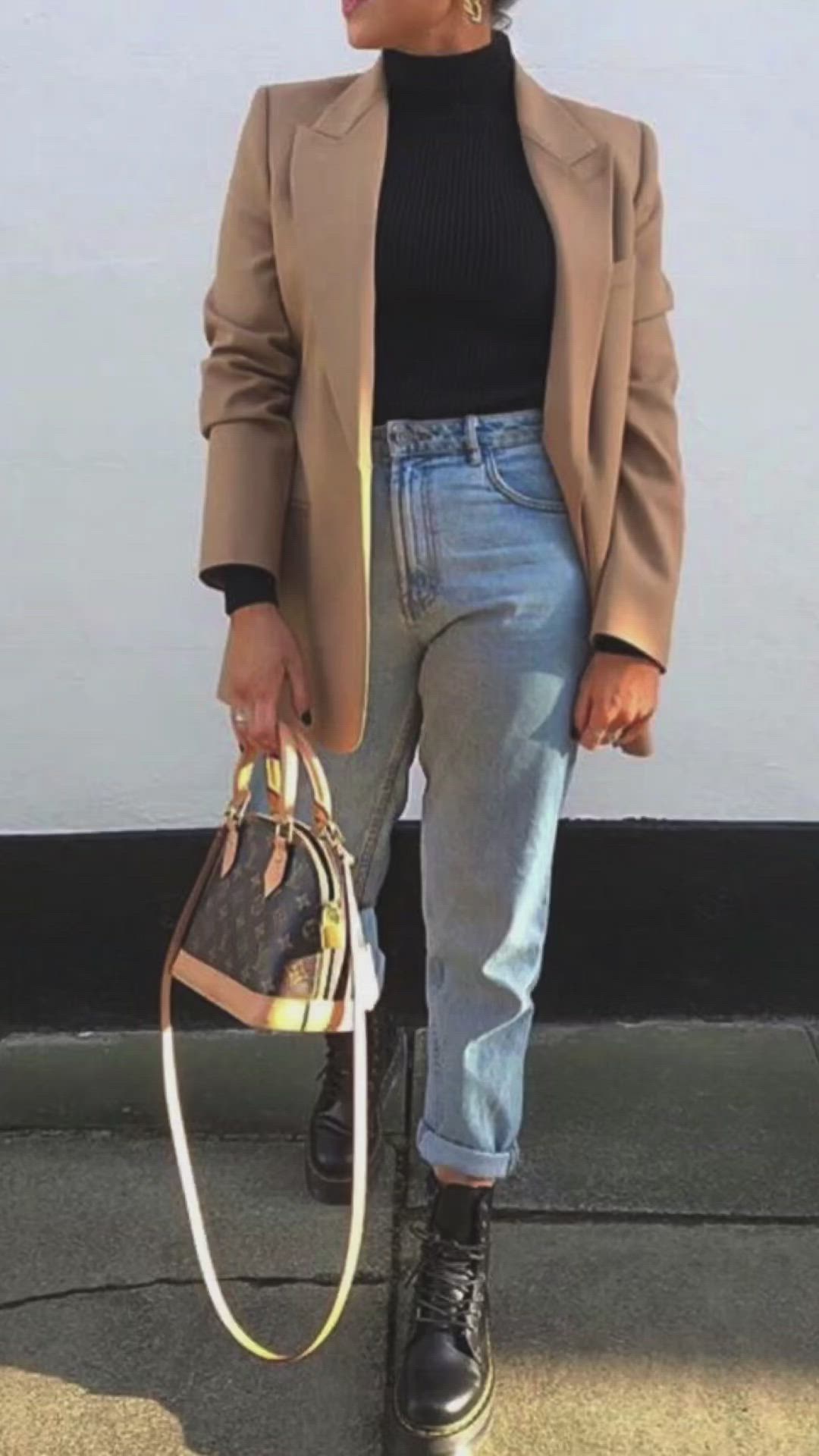 a woman is standing wearing a tan blazer, black turtleneck, blue jeans, and black boots. she is holding a louis vuitton bag in her left hand. she also appears to be wearing gold earrings.