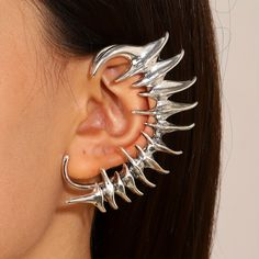 the image showcases a closeup view of a woman modeling a silver ear cuff. the ear cuff is designed with sharp, pointed edges, reminiscent of a dragon's spine or a mythical creature's claw.  it wraps around the curve of her ear, extending from the lobe to almost the top of the ear cartilage.