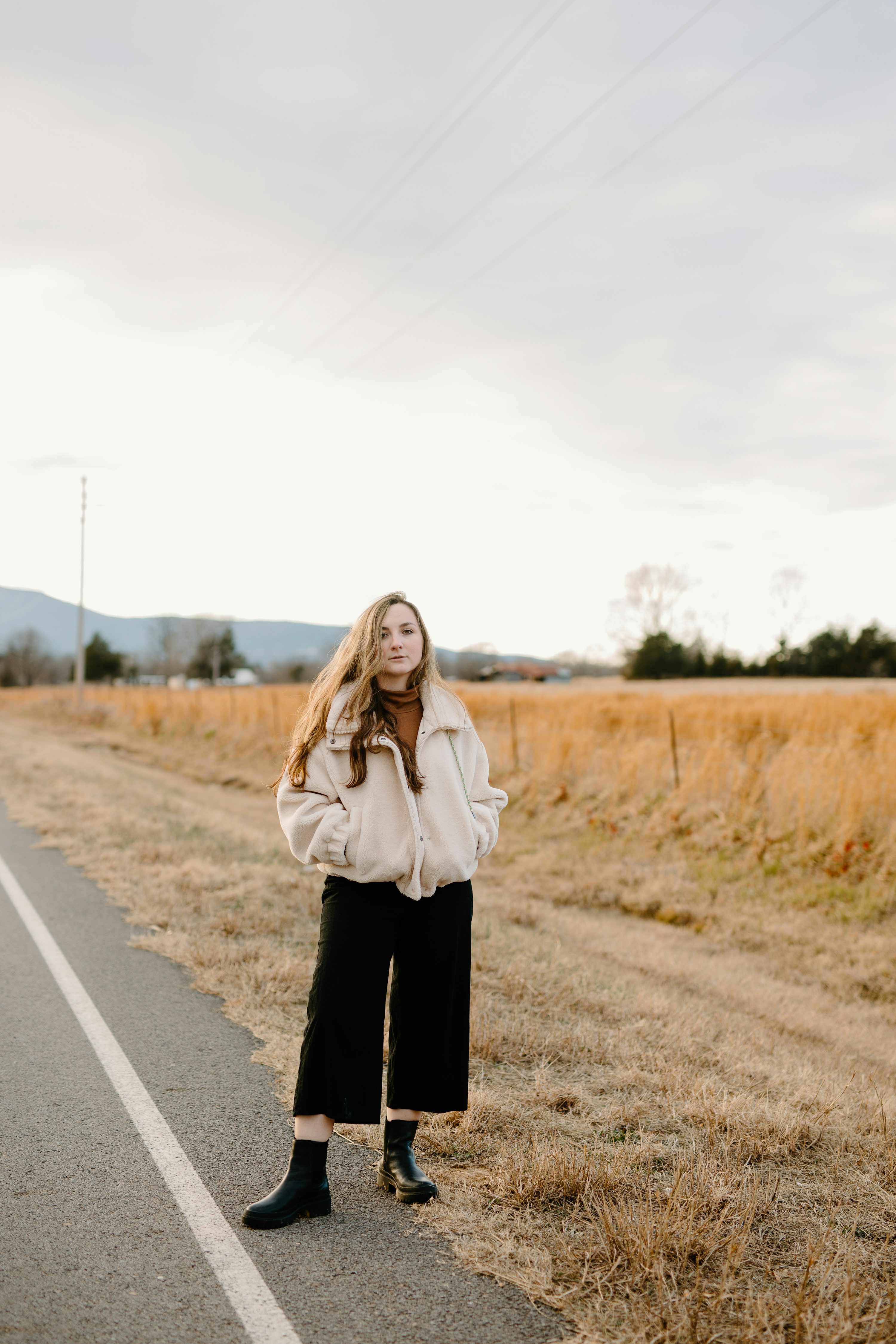 a young woman stands in the middle of a country road, wearing a creamcolored teddy bear jacket, black culottes, and black ankle boots. her long blonde hair flows freely. the road is bordered by dry, goldenbrown fields, and the sky is overcast. the overall atmosphere is calm and serene.