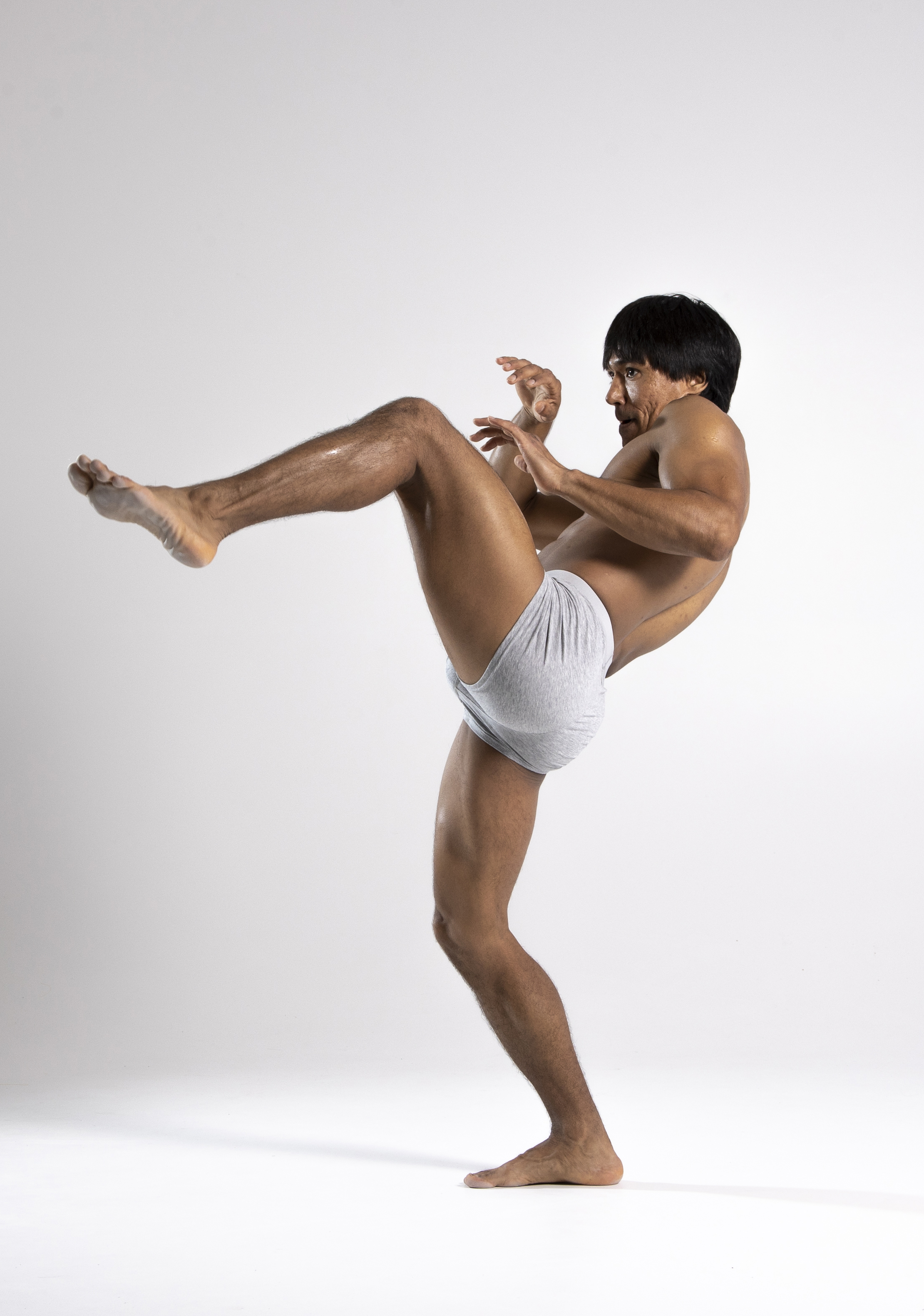 a shirtless man in grey underwear is captured midkick in a studio setting against a white background. he is barefoot, with his left leg raised high in a martial arts pose, displaying muscular legs and arms. his face shows concentration as his hands are held in a defensive stance. the dynamic pose suggests agility and athleticism.