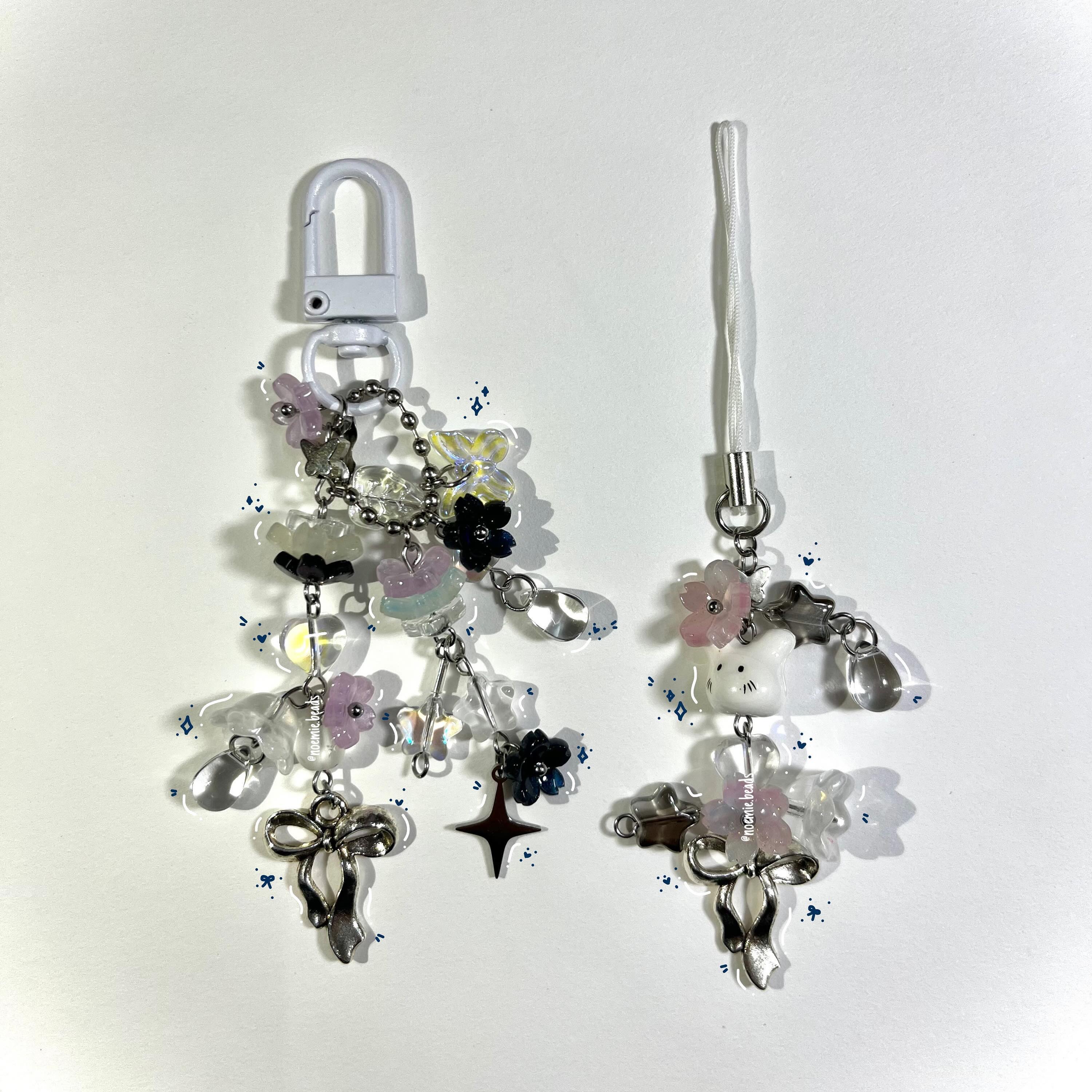 the image shows two handcrafted, beaded keychains against a white background. the keychain on the left has a white plastic clasp at the top followed by a chain of silver beads and a variety of charms, including clear resin shapes, lavender and black flowers, a silver bow, a black star, and clear teardropshaped beads. the keychain on the right has a white woven strap at the top connected to a series of charms, including pink flowers, a white cat face, clear resin shapes, and a silver bow. both keychains have a handmade, kawaii aesthetic with a mix of floral, pastel, and elegant elements. small blue sparkle graphics are added around the keychains.