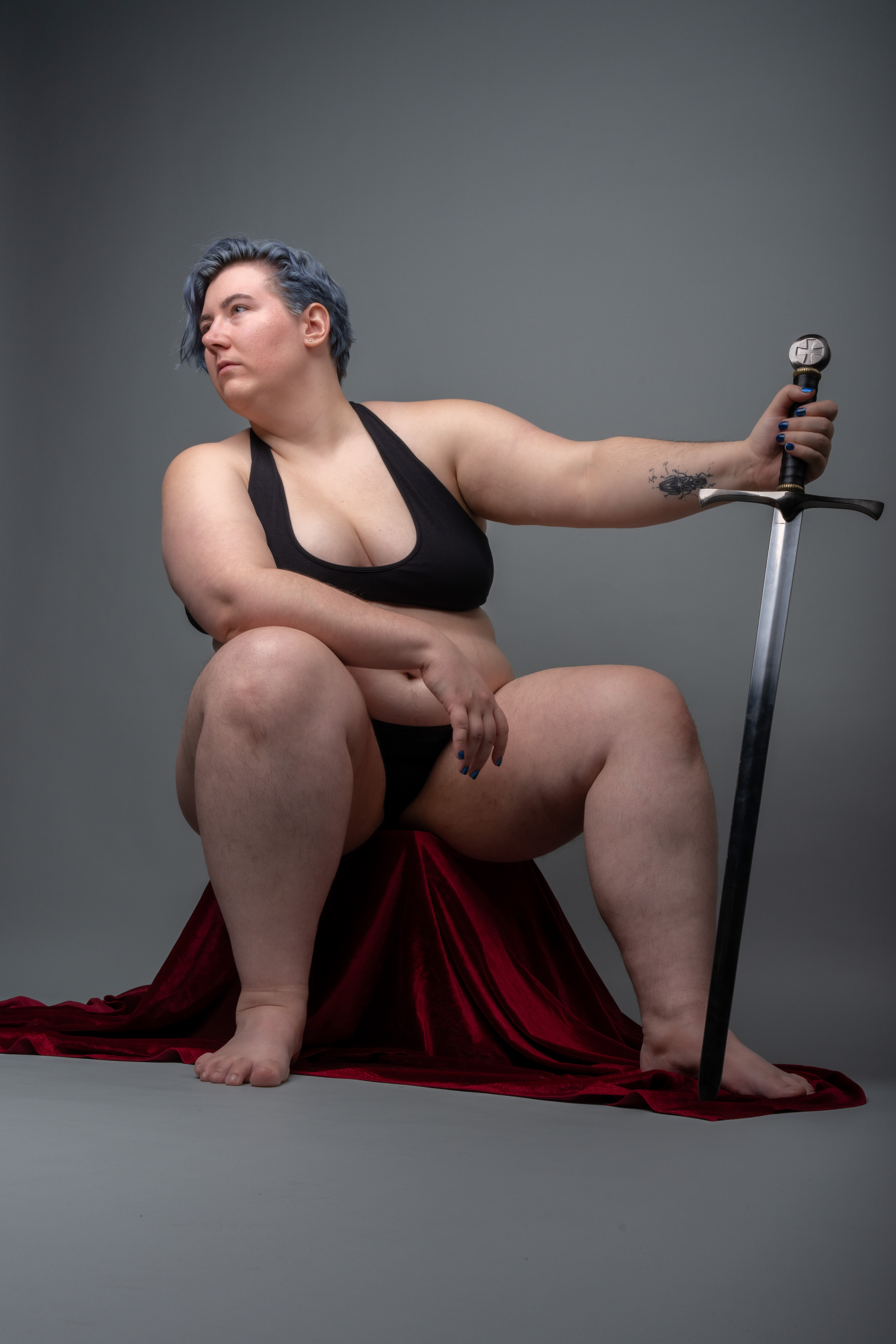 a studio shot of an overweight woman with blue hair sitting on a red cloth and holding a sword. she is wearing black underwear. the background is grey.
