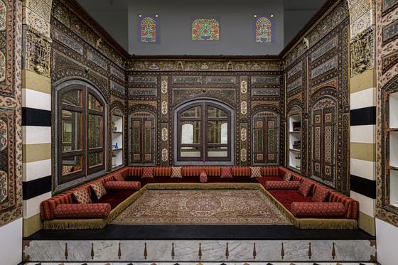 an elaborately decorated room featuring middle eastern design with intricate wood paneling, an oriental rug in the center, and luxurious cushions in a ushaped arrangement. the room has alcoves and windows, some with stained glass, all contributing to a traditional and vintage aesthetic.