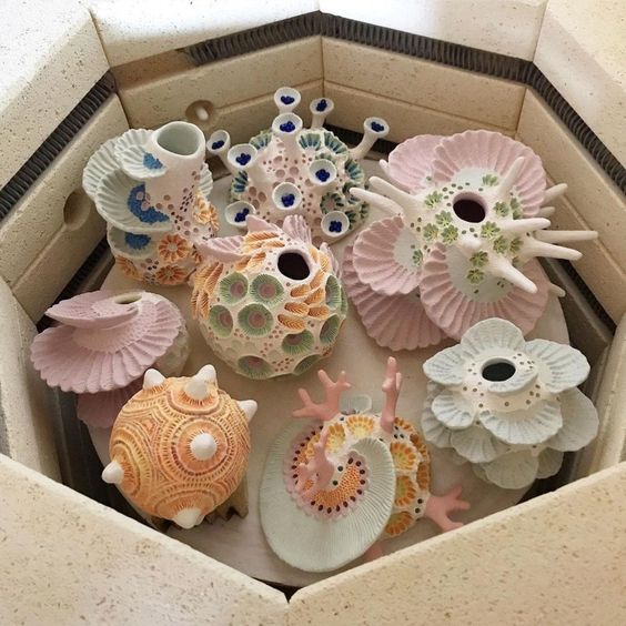 the image shows the inside of a kiln filled with colorful ceramic sculptures. the sculptures are arranged on a circular platform and are various shapes and sizes, many resembling coral or floral forms. they are predominantly pastel pink, blue, yellow, and green with intricate details and textures.