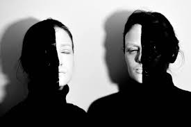 two women, with half of their faces painted black and half white, stand facing the camera. the woman on the left has a light background and the woman on the right has a dark background. both women are wearing black turtlenecks and looking at the camera. the image is in black and white and has a dramatic, mysterious feel to it.  it's a powerful portrait that plays with contrast and light to create a visual effect of duality or even opposites.
