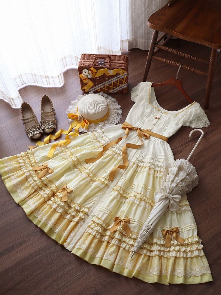 the image shows a pale yellow lolita fashion dress laid out on a wooden floor.  the dress is styled with a straw boater, brown mary jane shoes, a wicker handbag, and a white parasol. a wooden chair is visible in the top right corner.  the entire scene is brightly lit with natural light.