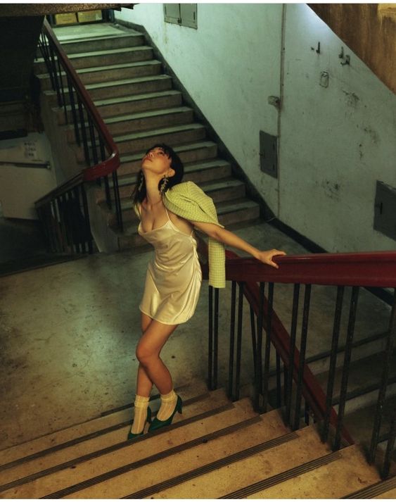 a woman in a white slip dress and green jacket is posing on a wooden staircase. she is looking up and her arm is resting on the railing. the walls and stairs are painted a light yellow and the railing is red. the lighting is artificial and the overall tone of the image is moody and atmospheric.