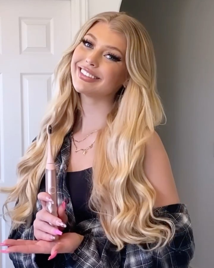 the image features loren gray, a wellknown influencer, smiling and holding an electric toothbrush. she has long, blonde hair styled in loose waves, and is wearing a black top with a plaid shirt draped over her shoulders. two delicate necklaces adorn her neck, and her nails are painted pink. the background includes a doorway and a glimpse of an interior space.