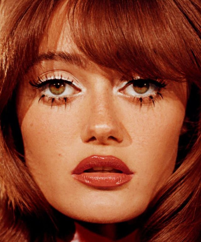 a closeup portrait of a woman with auburn hair, bangs, and prominent eye and lip makeup. the woman has fair skin and freckles. she is wearing heavy eye makeup with white eyeliner and long, unusual lower lashes. her lips are painted with glossy red lipstick. the overall style suggests a vintage or retro aesthetic.