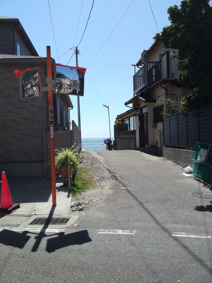 a street in japan, flanked by houses and leading to the sea, with street mirrors and a traffic cone visible.