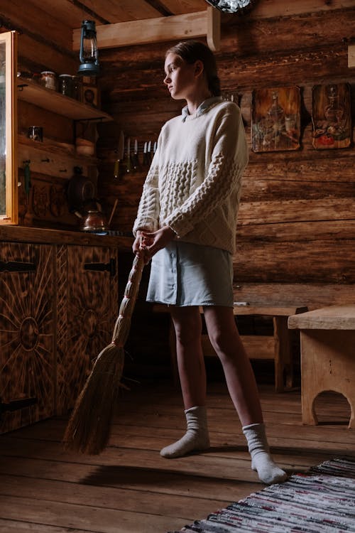 a young woman, possibly a teenager, stands in a rustic wooden kitchen holding a broom. she's wearing a creamcolored knitted sweater, a light blue skirt, and socks. the setting is a traditional log cabin with wooden walls, a wooden floor, and vintage kitchen utensils and furniture. the girl looks thoughtful and calm, creating a peaceful atmosphere. the image has a vintage and slightly melancholic feel.  the scene suggests a rural or country setting.