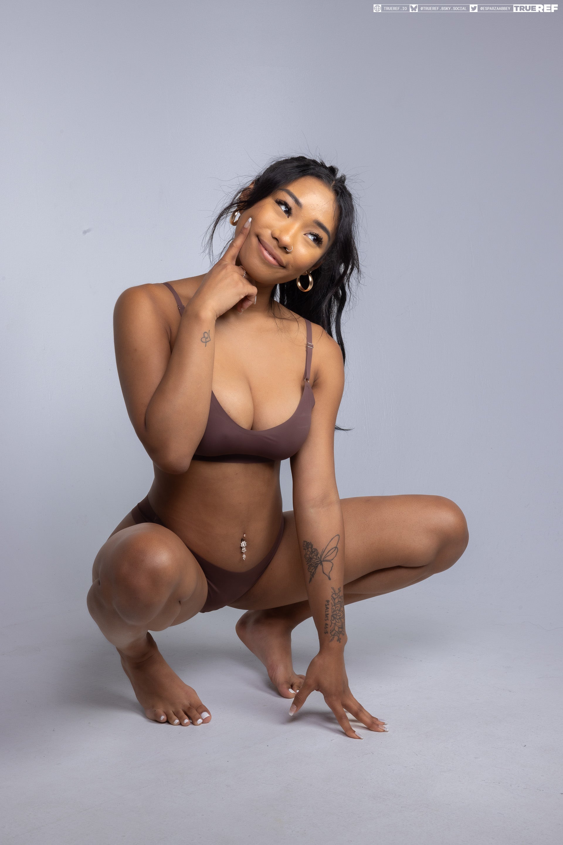 a young woman is crouching on the floor, looking up and to the right while touching her face with her finger. she is wearing a brown bra and underwear set. she has a tattoo on her arm and an earring. her navel is pierced. she is kneeling on the floor with her feet together. the background is a light gray color.