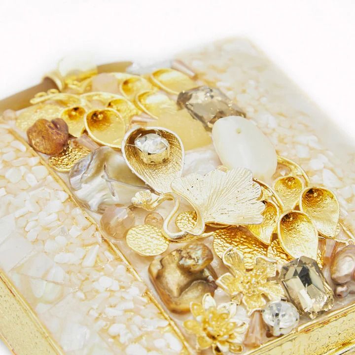 a closeup image features a decorative handbag. the handbag is adorned with various embellishments, including pearls, beads, gold accents, and other jewelry pieces. the overall aesthetic is ornate and luxurious, suggesting a highend fashion accessory.