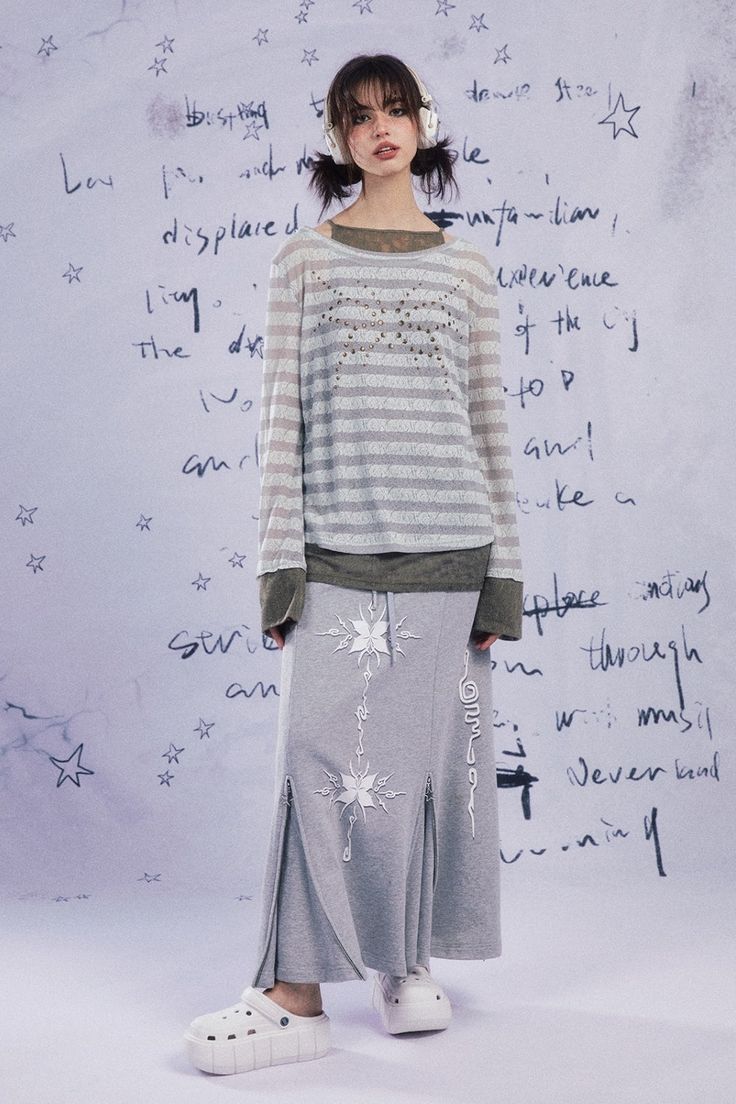 a fullbody shot of a young woman modeling a stylish outfit. she's wearing a longsleeved, light grey and white striped oversized sweater, layered over a dark olive green top. the sweater has a subtle, possibly embroidered, design on the chest. the skirt is a long, light grey piece with a drawstring waist, side slits, and a snowflakelike embroidery or applique pattern down the front. the model is also wearing white platform clogs. she has on overear headphones. the backdrop is a pale lilacgrey wall with handwritten text and star drawings scattered across it, adding a grunge/graffiti aesthetic to the image. the overall style of the photo is casual yet trendy, with a slightly grunge feel. the lighting is soft and diffused.
